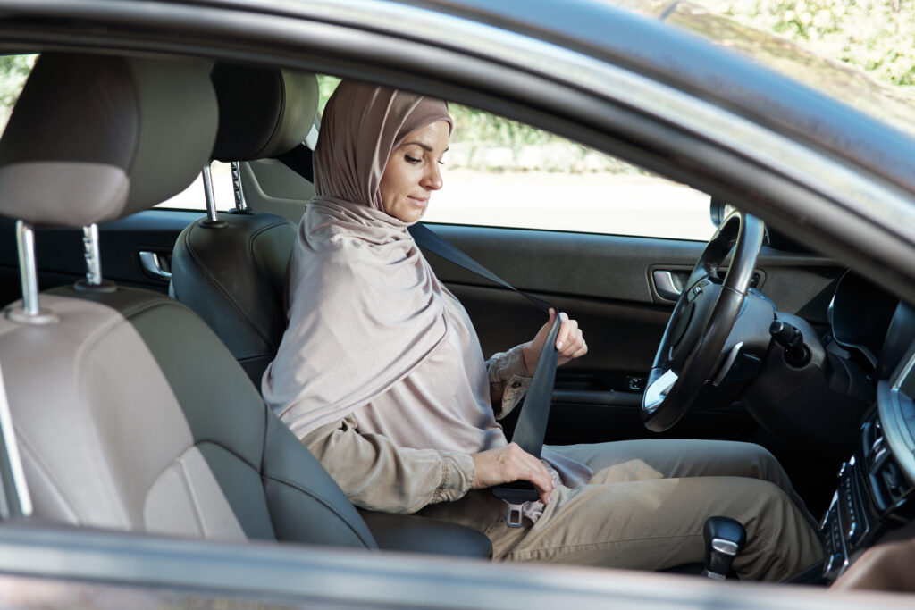 Horizontal shot of modern Muslim woman wearing hijab sitting in her car buckling seatbelt before starting driving. Don't drive when tired to stay safe when driving.